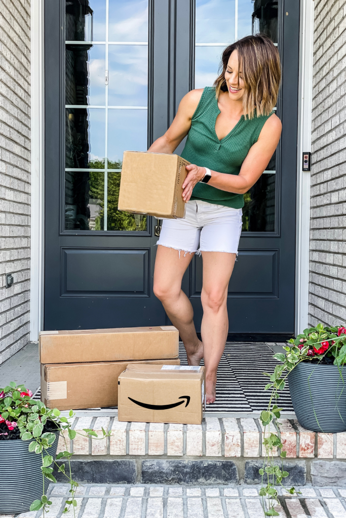 woman holding cardboard box with pile of more boxes at her feet on her front porch, one box is labeled as an Amazon box | Amazon Prime Day 2021 Finds on HauteHouseLove.com